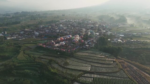 Aerial view of colorful hot air balloons being prepared for launch in a lively field near Wonosobo, with the majestic Mount Sindoro rising in the background.