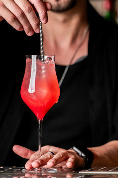 A bartender stiring a vibrant red cocktail in a long-stemmed tulip glass on a bar counter. Close-up shot with a blurred background