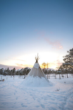 Traditional Sami lavvu tent standing in snowy Arctic landscape at sunrise. Warm light contrasts cold winter scene, capturing Nordic culture, wilderness and peaceful remote environment