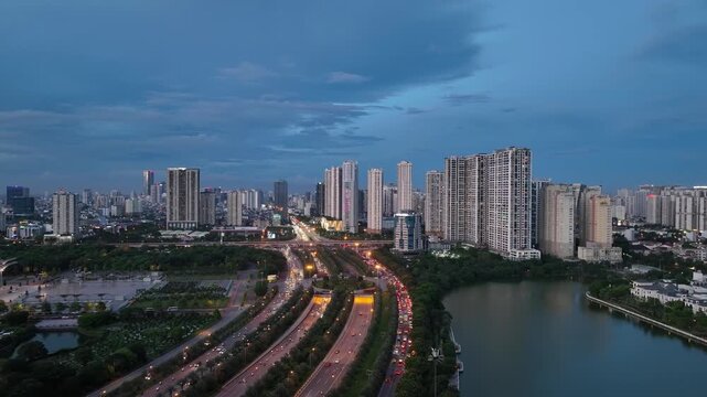 Drone perspective of curving Thang Long Boulevard during blue hour with heavy traffic flow, modern skyline, and lakeside residential buildings in Hanoi