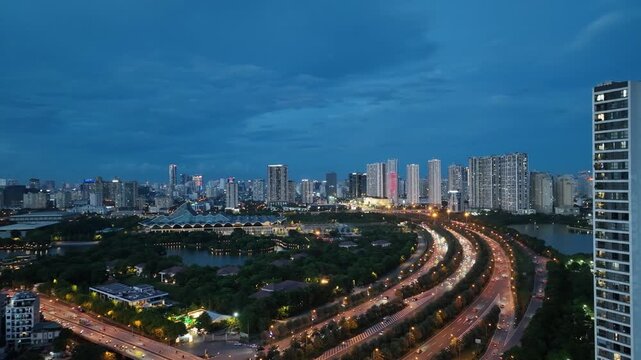 Blue hour drone scene of Thang Long Boulevard with flowing traffic, lakeside urban district, and illuminated skyline creating dynamic Hanoi atmosphere