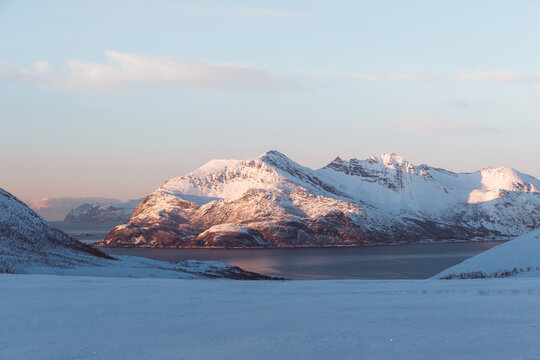 Golden sunset over a fjord on Kvaloya, Norway, with sun rays spreading above snow-covered mountains