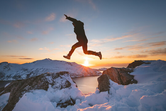 Silhouette of a person jumping between snowy rocks above a fjord on Kvaloya, Norway. Golden sunset light