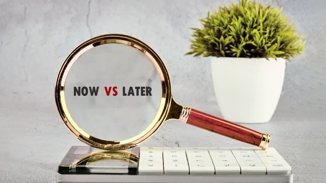 A golden magnifying glass highlights the words Now versus Later on a clean grey desktop, situated next to a white keyboard and a green potted plant