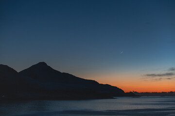 Fototapeta premium Twilight over a fjord in Norway with a crescent moon above the horizon. Calm Arctic waters, dark mountain