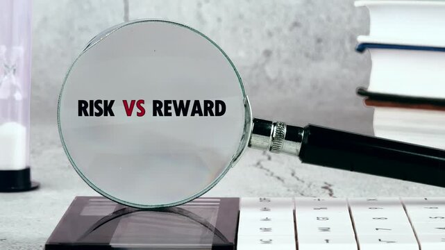 A magnifying glass reveals the words Risk vs Reward in clear focus. It rests on a light gray office desk with books, a pen, and an hourglass nearby