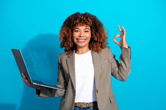 Young female business professional with curly hair holds a laptop and makes an ok gesture against blue background for lifestyle and office stock photo