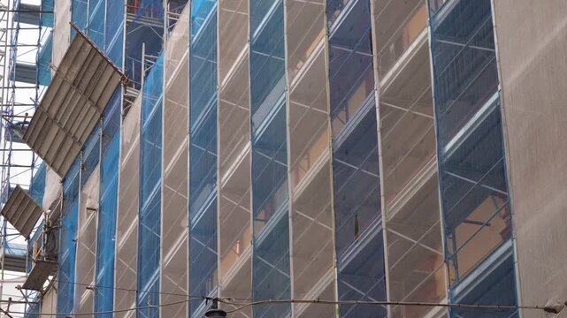 A modern construction hoist and scaffolding. The building is extensively clad in multi-level metal scaffolding covered with blue-beige protective mesh. Urban renovation, construction.