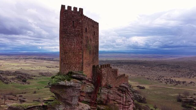 Medieval tower of Castle of Zafra rising from red rocky outcrop in Guadalajara, Spain, with vast arid Castilian landscape and dramatic cloudy sky