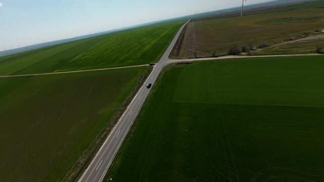FPV drone footage flying above vast green farmland, tracking a straight rural highway and intersection stretching to the horizon; minimalist aerial landscape under clear sky.