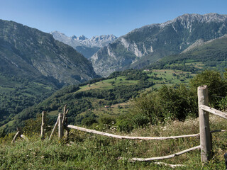 Fototapeta premium Mountain Valley View with Rustic Wooden Fence - Stunning panorama of a lush green mountain valley with a rustic fence and jagged peaks under a clear blue sky.