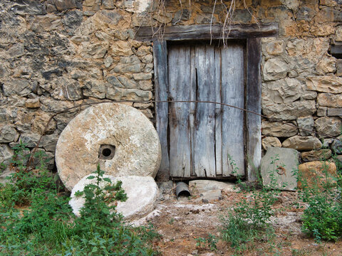 Ancient Stone House with Millstones and Doors - Abandoned rural house facade with rustic stone wall, weathered wooden doors and windows, and historic millstones in the foreground.