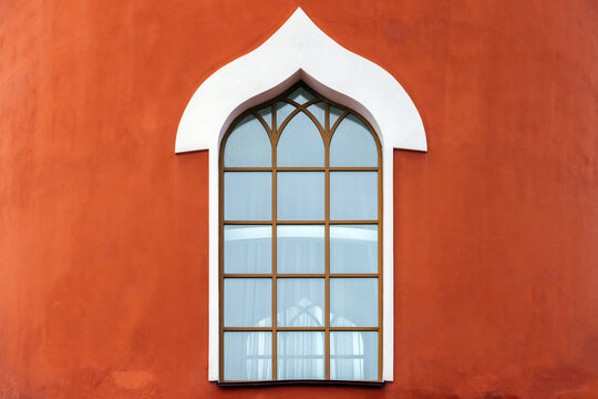 An ornate arched window with white trim set into a terracotta colored facade, featuring geometric mullions and soft blue glass that creates calm, minimalist architectural composition. Background photo