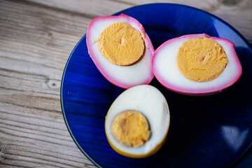 Close-up of halved pickled eggs with vibrant pink and yellow whites on a dark blue plate, showing firm yolks and shallow depth of field. Soft background blur. © Jolanta