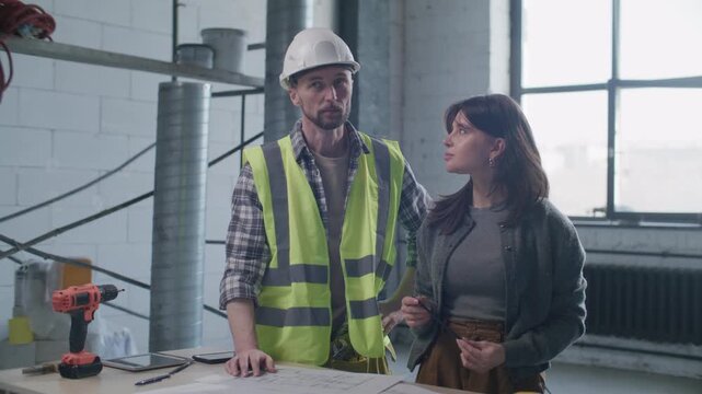 Foreman in safety vest and hardhat reviewing floor plan with female client at construction site, discussing project details and planning workflow