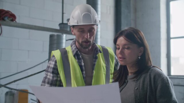 Industrial engineer in hard hat and safety vest explaining floor plan to female client, pointing at blueprint and discussing project details with her at construction site