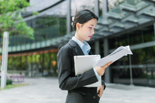 Asian young professional businesswoman in a black suit reads documents outdoors near a modern office building, holding a tablet under her arm while focused on her work.