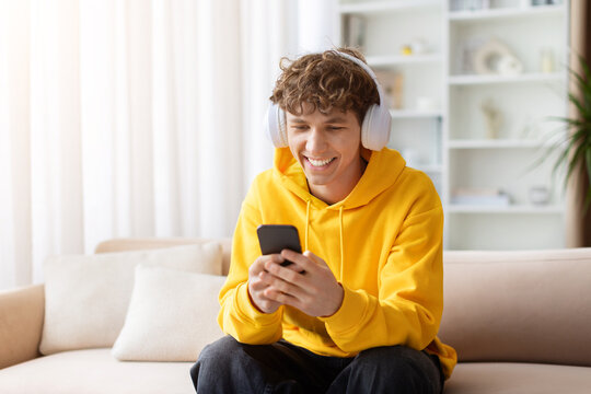Young smiling teen guy with white wireless headphones sitting on sofa, looking at smartphone. Happy male teenager in yellow hoodie using phone at home