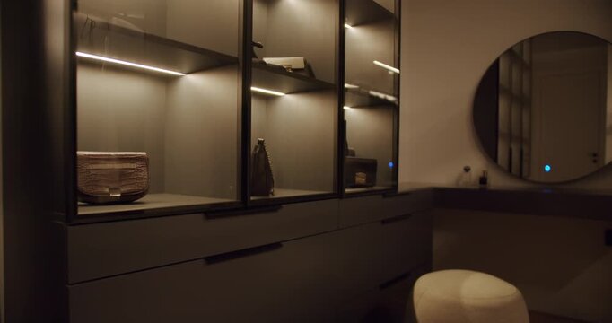 Modern walk-in closet with illuminated glass shelves and drawers. Stylish handbags and accessories displayed. Round mirror and vanity area. Elegant interior design. Luxury apartment living.