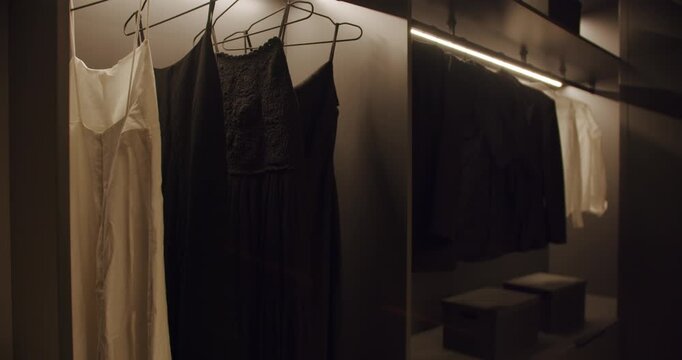Modern walk-in closet with elegant clothes and soft lighting. Dark wardrobe with illuminated shelves and hanging garments. Stylish interior design for fashion and storage.