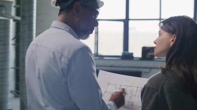 Female homeowner reviewing and discussing floor plan with senior male contractor in hard hat during consultation on construction site. Back view