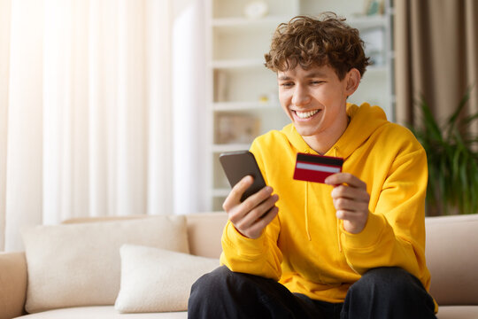 Smiling teen boy in yellow hoodie holding credit card and smartphone, making mobile payment on sofa. Happy young male shopping online at home