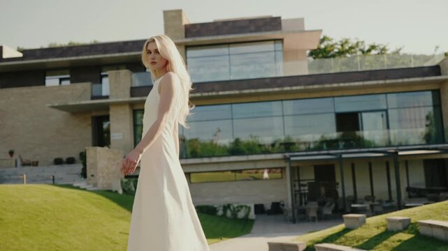 A young woman in a flowing white dress walks outside a contemporary luxury house. Elegance, modernity, and sophistication are exuded in this serene outdoor setting. Elegant Woman in White Dress