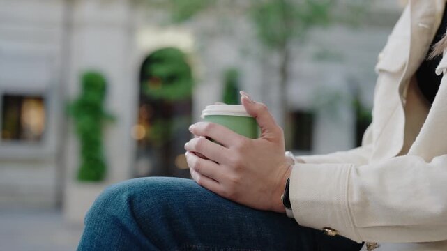 Close-up of a woman holding a green coffee cup while sitting outdoors. The scene is calm, highlighting a relaxed moment in an urban environment with greenery in the background.