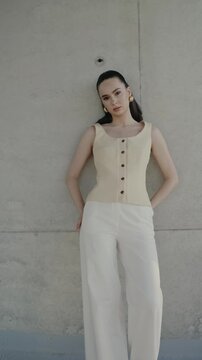 Elegant young woman posing against a concrete wall, wearing a stylish sleeveless top and white pants. Fashion, style, and confidence are prominent in this minimalist outdoor shot.