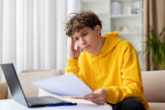 Worried teen boy in yellow hoodie reading paper document at laptop with hand on temple, looking stressed. Young troubled male reviewing paperwork at home