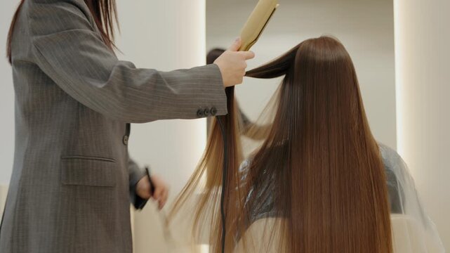 A professional hairdresser uses a hair straightener to style a client's long, brown hair in a modern salon. The stylist works meticulously, section by section, to create a sleek and polished look.