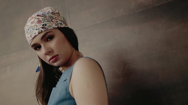 Portrait of a young woman with a serious expression, wearing a stylish bandana and blue top. Neutral background adds focus to her face and attire, giving a modern and contemplative feel.