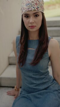 A woman sits on steps, wearing a floral headscarf and a blue dress, looking directly at the camera with a calm expression. Outdoors setting. Young Woman in Blue Dress with Headscarf, Outdoor Portrait