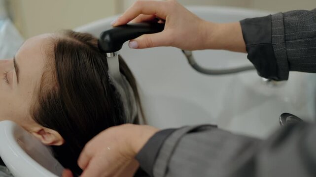 A woman gets her hair washed at a hair salon. The stylist gently rinses her client's hair with water. The client has her eyes closed and appears relaxed.