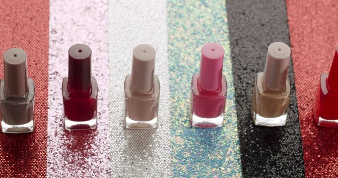 Assortment of colorful nail polish bottles arranged in a row over a shiny, multicolored glitter background, showing a palette of beauty products for manicure and pedicure in slow motion