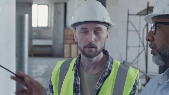 Site foreman in safety gear standing inside unfinished building, pointing at architectural schemes on wall and explaining project details to senior engineer