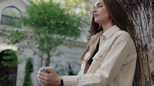 A woman in casual clothing leans against a tree, enjoying a peaceful moment outdoors. The background shows a building with greenery. Woman Enjoying a Peaceful Moment Outdoors Leaning Against a Tree