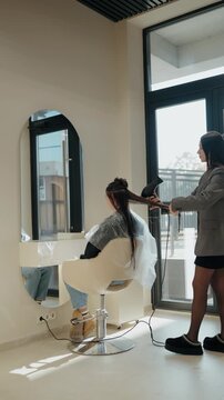 A hair stylist blow-dries a client's long brown hair in a salon. The client sits in a chair and looks in the mirror, which is an oval shape. The stylist uses a black hairdryer to dry the client's hair