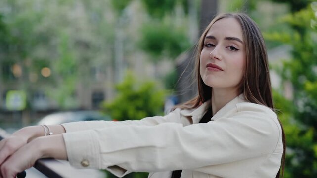 Portrait of a young woman wearing a beige jacket, leaning on a railing while enjoying time outdoors. The background features lush greenery, enhancing a relaxed and confident vibe.Woman Posing Outdoors