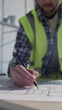 Professional builder in safety vest marking up technical drawings with pencil on construction site. Vertical shot, close-up view