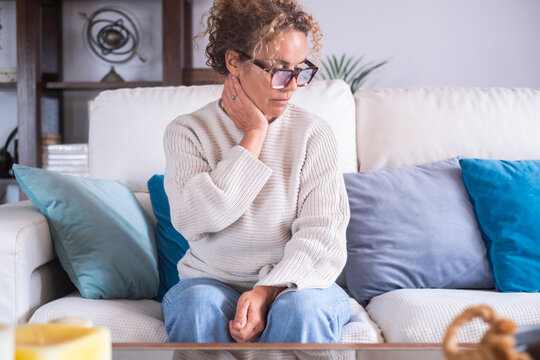 Mature woman sitting on sofa holding neck in pain at home looking concerned and thoughtful