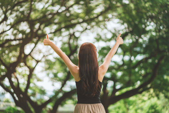 Back view of a young woman with headphones raising her thumbs up in a lush green park, celebrating success and freedom through music and nature during a sunny day to promote mental wellness and a posi