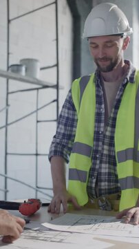 Smiling construction worker in hard hat and safety vest giving handshake to site manager over table with floor plans, power drill and notebook inside unfinished building. Vertical clip