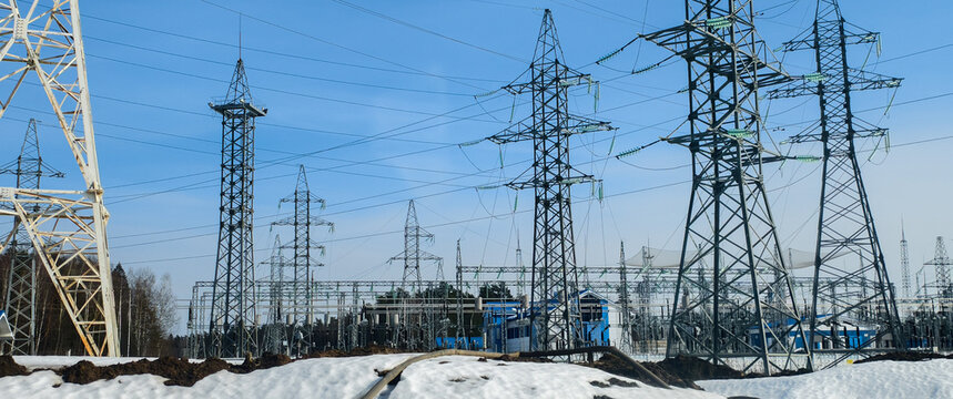 Power lines and electrical substation featuring several high-voltage transmission towers and conductors against a clear blue sky, demonstrating an energy infrastructure network on a snowy landscape.