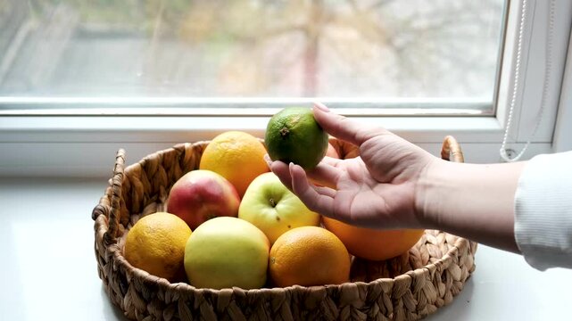 Woven basket on windowsill holds grapefruit, apples, orange, lime. Girl touches and inspects fruits. Early spring outdoors, reminder to get enough vitamins. 