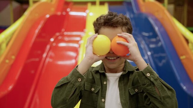 Happy cute little African American boy posing in the game center while covering his eyes with balls, slide on the background. Slow motion