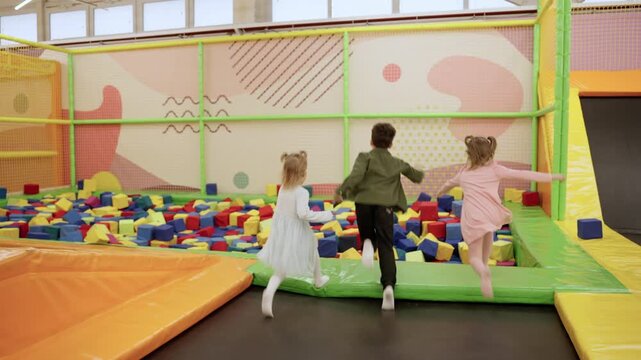 Two little girls and boy playing on indoor playground in foam rubber pit in trampoline, running and jumping. Slow motion