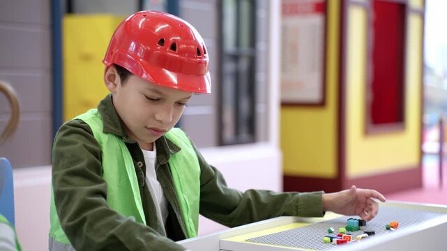 Little African American boy in helmet and waistcoat playing with colorful toy blocks, building tower in game center. Slow motion
