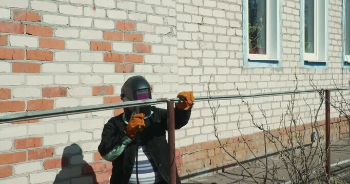Skilled male worker in a protective mask performing electric welding on a metal fence structure against a brick wall, with bright sparks flying from the hot metal during the fabrication process.