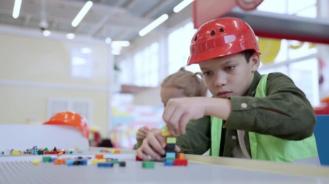 Little African American boy in safe helmet building tower using colorful toy blocks on the table in educational gaming center. Slow motion